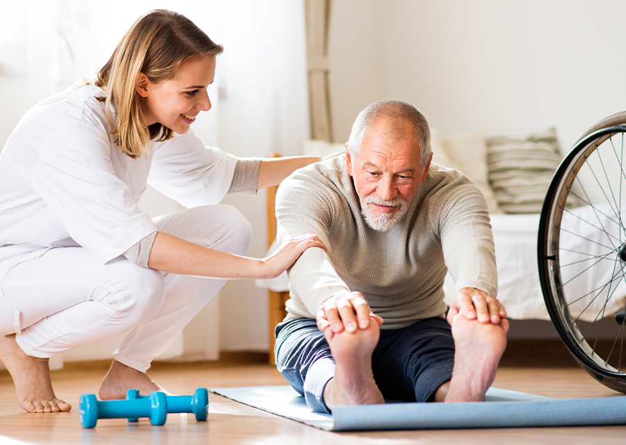 Senior sitting on a yoga mat grabs his toes and stretches with the assistance of a physical therapist