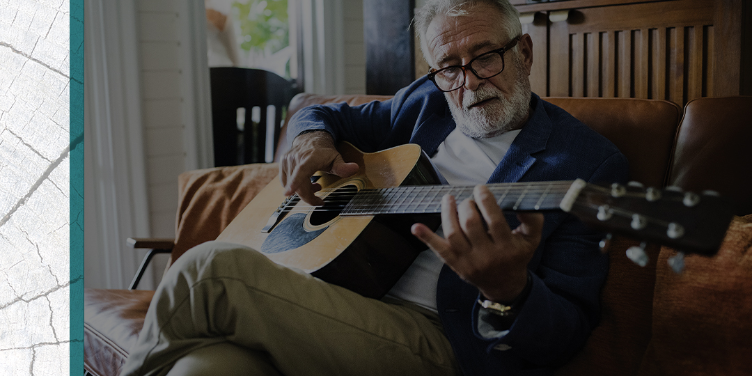 Smartly dressed bearded senior plays guitar on a couch.